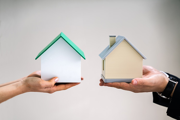 Close-up,Of,Couple's,Hand,Holding,Different,Miniature,Houses,Against,Gray